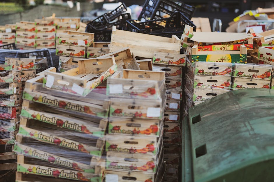 A large outdoor area filled with numerous wooden and plastic crates, predominantly decorated with images and branding of fresh fruits, stacked haphazardly. The wooden crates show signs of weathering, 