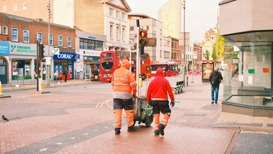 Two waste collection workers dressed in high-visibility orange and red jackets with reflective strips are pushing a wheeled bin along a city sidewalk. The workers are seen from behind, with one turnin