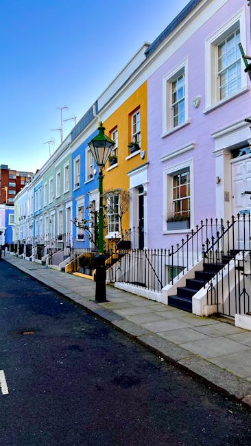 A row of terraced houses painted in pastel shades, including light purple, yellow, and blue, lining a street with a paved sidewalk. Each property features steps leading up to white-fronted doors, blac