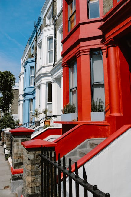 A row of terraced houses painted in pastel shades, including light purple, yellow, and blue, lining a street with a paved sidewalk. Each property features steps leading up to white-fronted doors, blac