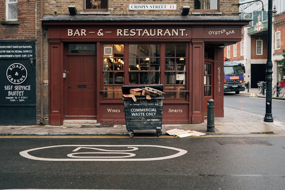 In front of a red-brick building housing a bar and restaurant with large front windows and a wooden exterior, a black commercial waste bin labeled 'COMMERCIAL WASTE ONLY' is positioned on the sidewalk