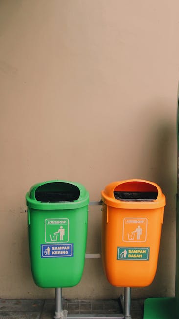 A person in a green winter coat, striped hat, and dark trousers is seen standing beside four large green communal wheelie bins inside a covered metal shelter on a snowy street. The bins are filled wit