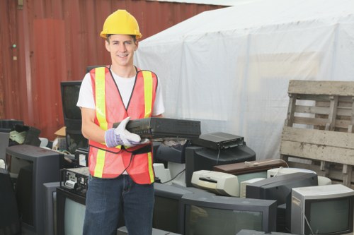A young man wearing a yellow helmet, white T-shirt, and high-visibility safety vest holding a black electronic device while standing outdoors in front of a collection of discarded old televisions and monitors. The background features a red fence and a white tarp or tent, suggesting an outdoor waste collection or rubbish clearance scenario. The scene indicates a focus on electronic waste removal, with the individual appearing to be a worker or volunteer engaged in rubbish clearance activities, relevant to services offered by Garden Clearance London within the London area, possibly near the postcode inferred from the webpage. The environment is well-lit, with a casual atmosphere typical of a waste disposal or rubbish removal site, emphasizing the handling of bulky or electronic waste to support rubbish clearance services in the London region.