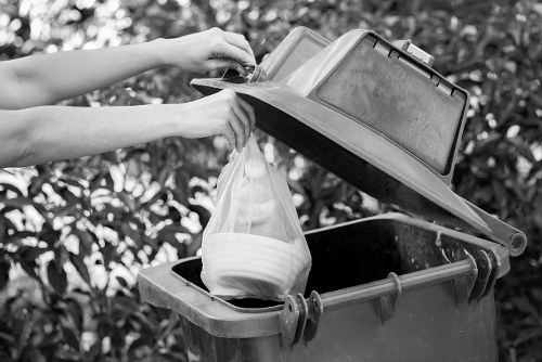 A person’s hands are shown disposing of a clear plastic bag filled with white ceramic bowls into a black rubbish bin with a hinged lid, situated outdoors on a paved surface with foliage in the background. The lid of the bin is open, revealing the interior, and the person appears to be carefully placing the bag inside. The scene reflects common rubbish disposal activity, which could relate to garden clearance or general waste collection in the London area. The bin is positioned on a driveway or pathway, with the lid tilted back, and the environment suggests a domestic setting suitable for rubbish removal services provided by companies such as Garden Clearance London. Natural light highlights the textures of the plastic bag, ceramic material inside, and the surfaces of the bin. The overall scene emphasizes typical waste disposal tasks associated with garden clearance or home rubbish collection in the London region, relevant to the service category of rubbish removal.